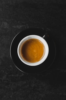 A cup of coffee served in a white cup with a black saucer, placed against a dark textured background. The coffee appears rich and smooth, with a layer of crema on top.