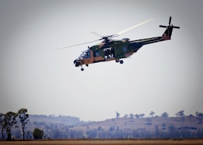 A helicopter is flying above a rural landscape with some trees and hills visible in the background. The helicopter is green with some camouflage patterns and has its side doors open.