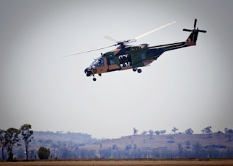 A helicopter is flying above a rural landscape with some trees and hills visible in the background. The helicopter is green with some camouflage patterns and has its side doors open.