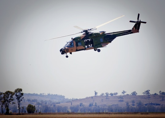 A helicopter is flying above a rural landscape with some trees and hills visible in the background. The helicopter is green with some camouflage patterns and has its side doors open.