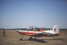 A small, single-engine aircraft with a distinctive orange and white color scheme is parked on a grassy field under a clear blue sky. The aircraft is a propeller type and appears to be used for training or basic flight exercises. A person wearing a uniform is seen standing on the field, seemingly observing or preparing the aircraft.
