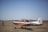 A small, single-engine aircraft with a distinctive orange and white color scheme is parked on a grassy field under a clear blue sky. The aircraft is a propeller type and appears to be used for training or basic flight exercises. A person wearing a uniform is seen standing on the field, seemingly observing or preparing the aircraft.