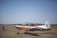 A small, single-engine aircraft with a distinctive orange and white color scheme is parked on a grassy field under a clear blue sky. The aircraft is a propeller type and appears to be used for training or basic flight exercises. A person wearing a uniform is seen standing on the field, seemingly observing or preparing the aircraft.