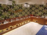 A buffet setup with a series of metal serving dishes arranged on a wooden counter. The background features vibrant green jungle-themed wallpaper with large leaf patterns. The ceiling has air conditioning units and a modern chandelier. The floor includes a geometric patterned tile design. Golden decorative objects are placed on lower shelves.
