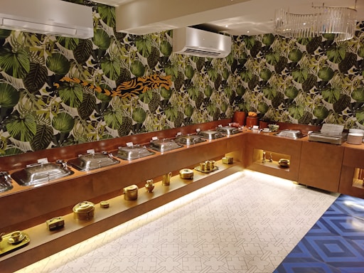A buffet setup with a series of metal serving dishes arranged on a wooden counter. The background features vibrant green jungle-themed wallpaper with large leaf patterns. The ceiling has air conditioning units and a modern chandelier. The floor includes a geometric patterned tile design. Golden decorative objects are placed on lower shelves.