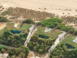 Aerial view of several small buildings with green roofs, surrounded by lush vegetation. The structures appear to be nestled close to a sandy beach, with a backdrop of natural shrubbery and expanses of sand.