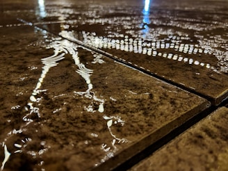 Water is pooling on tiled pavement, reflecting lights from above. The surface is slightly wet, creating a shimmering effect with reflections that appear streaked across the tiles. The focus is on the texture and the interplay of light and water.