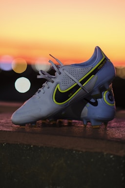 A pair of white soccer cleats with yellow and black detailing is propped on a surface against a backdrop of a vibrant sunset. The cleats are well-lit, showcasing their design and brand logo prominently.