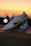 A pair of white soccer cleats with yellow and black detailing is propped on a surface against a backdrop of a vibrant sunset. The cleats are well-lit, showcasing their design and brand logo prominently.