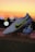 A pair of white soccer cleats with yellow and black detailing is propped on a surface against a backdrop of a vibrant sunset. The cleats are well-lit, showcasing their design and brand logo prominently.