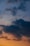 Dark, billowing clouds contrast against a gradually changing sky, transitioning from light blues at the top to warm shades of orange near the horizon.