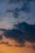 Dark, billowing clouds contrast against a gradually changing sky, transitioning from light blues at the top to warm shades of orange near the horizon.