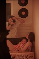 A man holding a camera is capturing a moment of a woman cradling a baby dressed in red and white stripes. The setting includes a home interior with two vinyl records on the wall and a warm, cozy ambiance.