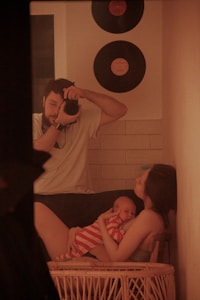 A man holding a camera is capturing a moment of a woman cradling a baby dressed in red and white stripes. The setting includes a home interior with two vinyl records on the wall and a warm, cozy ambiance.