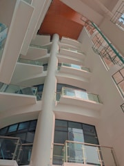 An interior view of a modern multi-story building with several floors visible from a low angle. Each floor has glass railings and windows, with a column running vertically in the center. The ceiling is covered in orange panels, and a green metal structure is seen on the right.