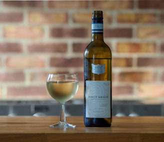A bottle of Pinot Grigio white wine and a filled wine glass are placed on a wooden surface with a blurred brick wall background. The wine bottle has a detailed label and a blue neck band.