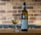 A bottle of Pinot Grigio white wine and a filled wine glass are placed on a wooden surface with a blurred brick wall background. The wine bottle has a detailed label and a blue neck band.