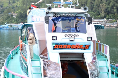 A colorful passenger boat with a unique design is docked by a calm, green lake. The boat displays vibrant branding on the front, and there are several figures on the upper deck. A person wearing a headscarf is standing on the side, looking out at the water. The surrounding area features lush greenery and buildings along the waterline.