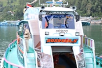 A colorful passenger boat with a unique design is docked by a calm, green lake. The boat displays vibrant branding on the front, and there are several figures on the upper deck. A person wearing a headscarf is standing on the side, looking out at the water. The surrounding area features lush greenery and buildings along the waterline.