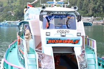 A colorful passenger boat with a unique design is docked by a calm, green lake. The boat displays vibrant branding on the front, and there are several figures on the upper deck. A person wearing a headscarf is standing on the side, looking out at the water. The surrounding area features lush greenery and buildings along the waterline.