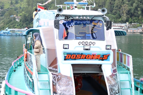 A colorful passenger boat with a unique design is docked by a calm, green lake. The boat displays vibrant branding on the front, and there are several figures on the upper deck. A person wearing a headscarf is standing on the side, looking out at the water. The surrounding area features lush greenery and buildings along the waterline.