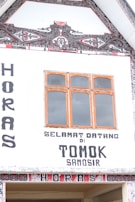 A traditional Batak-style building facade with intricate black and red geometric and floral patterns. The facade has large lettering welcoming visitors to Tomok, Samosir, with decorative woodwork around the windows.