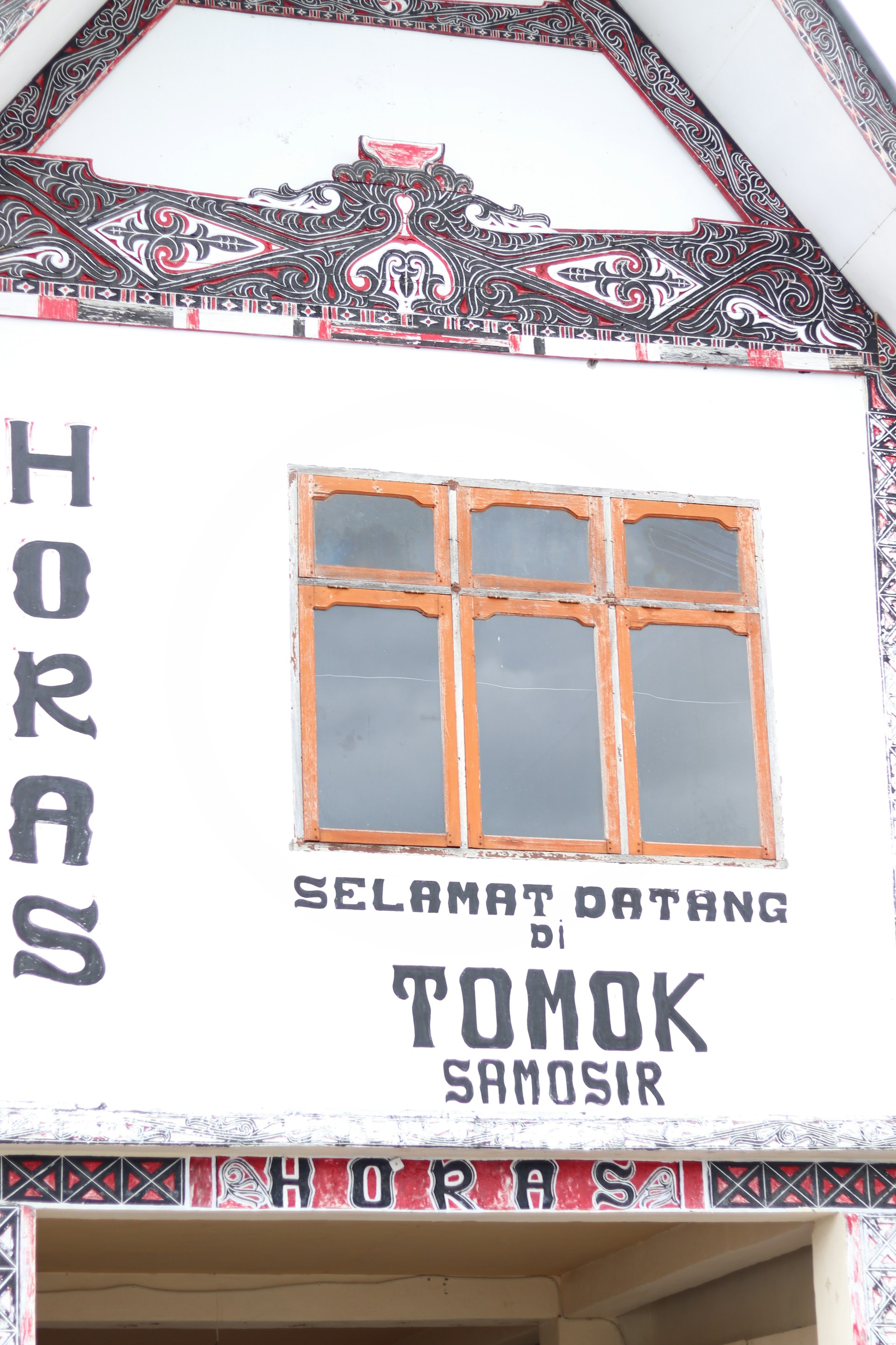 A traditional Batak-style building facade with intricate black and red geometric and floral patterns. The facade has large lettering welcoming visitors to Tomok, Samosir, with decorative woodwork around the windows.