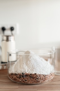 A clear glass container holds a layered mixture of white flour on top of a coarse blend of seeds and grains. The background features a blurred kitchen setting with minimalistic decor.