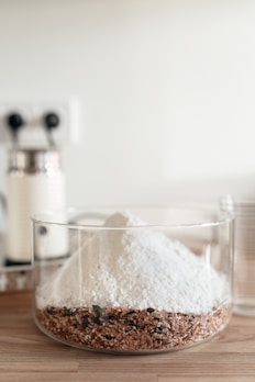 A clear glass container holds a layered mixture of white flour on top of a coarse blend of seeds and grains. The background features a blurred kitchen setting with minimalistic decor.