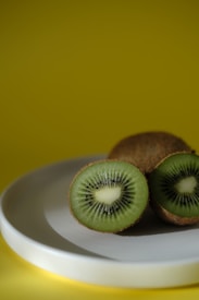 A close-up of a kiwi fruit cut in half displayed on a smooth white plate with a vibrant yellow background. The green flesh and tiny black seeds are prominent.