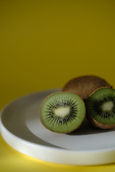 A close-up of a kiwi fruit cut in half displayed on a smooth white plate with a vibrant yellow background. The green flesh and tiny black seeds are prominent.