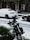 Snow covers a parked white car and a green motorcycle on a street. Snowflakes are visible throughout, indicating an ongoing snowfall. The background shows a building with a tiled facade and a large air conditioning unit attached.