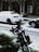 Snow covers a parked white car and a green motorcycle on a street. Snowflakes are visible throughout, indicating an ongoing snowfall. The background shows a building with a tiled facade and a large air conditioning unit attached.