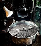 A large metal roasting machine contains a substantial amount of freshly roasted coffee beans. The beans are dark and glossy, filling the bottom of the circular roasting drum. An industrial-style lamp illuminates part of the machine, creating a contrast between light and shadow. The surrounding environment suggests an industrial or artisanal processing space, with the machine dominating the foreground.