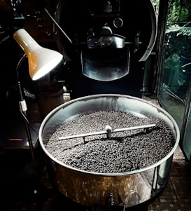A large metal roasting machine contains a substantial amount of freshly roasted coffee beans. The beans are dark and glossy, filling the bottom of the circular roasting drum. An industrial-style lamp illuminates part of the machine, creating a contrast between light and shadow. The surrounding environment suggests an industrial or artisanal processing space, with the machine dominating the foreground.