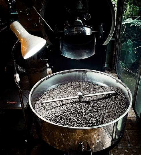 A large metal roasting machine contains a substantial amount of freshly roasted coffee beans. The beans are dark and glossy, filling the bottom of the circular roasting drum. An industrial-style lamp illuminates part of the machine, creating a contrast between light and shadow. The surrounding environment suggests an industrial or artisanal processing space, with the machine dominating the foreground.