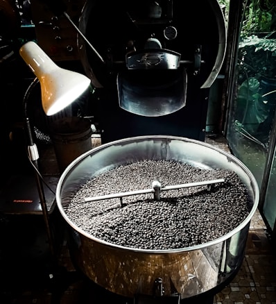 A large metal roasting machine contains a substantial amount of freshly roasted coffee beans. The beans are dark and glossy, filling the bottom of the circular roasting drum. An industrial-style lamp illuminates part of the machine, creating a contrast between light and shadow. The surrounding environment suggests an industrial or artisanal processing space, with the machine dominating the foreground.