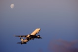 A commercial airplane is captured mid-flight against a clear sky with the moon visible in the background. The aircraft's branding is discernible, and the evening light casts a warm glow on its fuselage.