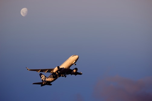 A commercial airplane is captured mid-flight against a clear sky with the moon visible in the background. The aircraft's branding is discernible, and the evening light casts a warm glow on its fuselage.