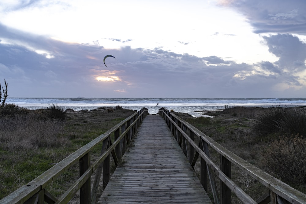 a wooden walkway leading to a beach with a kite flying in the sky