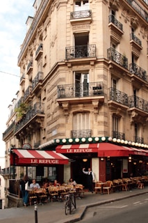 A charming street corner cafe with red and white striped awnings, situated in a historic European building with ornate wrought iron balconies and detailed stonework. Several people are seated outside the cafe enjoying drinks and conversation, while a server stands at the entrance.