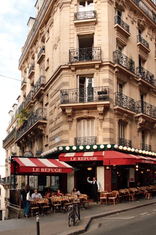 A charming street corner cafe with red and white striped awnings, situated in a historic European building with ornate wrought iron balconies and detailed stonework. Several people are seated outside the cafe enjoying drinks and conversation, while a server stands at the entrance.