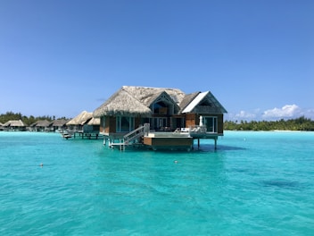A serene overwater bungalow with a thatched roof is set against a backdrop of crystal-clear turquoise waters and a clear blue sky. The bungalow features glass windows and steps leading down to the water. In the distance, additional bungalows can be seen, and lush green foliage lines the horizon.