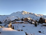 A snowy alpine village nestled at the base of a mountain range bathed in sunlight. The scene is dominated by wooden chalets and buildings with snow-covered roofs, surrounded by evergreen trees. People are skiing and walking in the foreground, indicating a lively atmosphere in a ski resort setting.