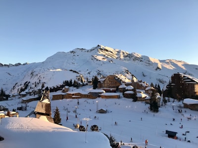 A snowy alpine village nestled at the base of a mountain range bathed in sunlight. The scene is dominated by wooden chalets and buildings with snow-covered roofs, surrounded by evergreen trees. People are skiing and walking in the foreground, indicating a lively atmosphere in a ski resort setting.