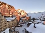 A snowy alpine village at sunset with wooden chalet-style buildings lining the street. The snow-covered mountains in the background are lit with a warm glow from the setting sun. People are seen walking along the main street, enjoying the winter atmosphere.
