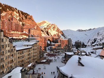A snowy alpine village at sunset with wooden chalet-style buildings lining the street. The snow-covered mountains in the background are lit with a warm glow from the setting sun. People are seen walking along the main street, enjoying the winter atmosphere.