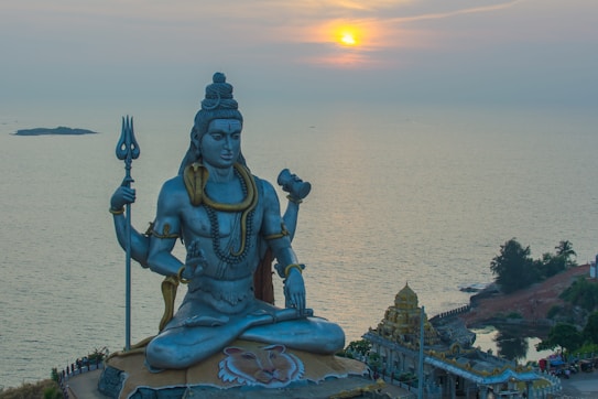 A large statue of a Hindu deity sits majestically by the sea during sunset. The figure is adorned with intricate jewelry and is holding a trident. The serene expression on the statue's face overlooks the calm waters and distant horizon. Lush greenery and temple-like structures are visible nearby.