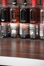 Several bottles of Sunich flavored syrups are lined up on a wooden surface. The flavors visible include chocolate and hazelnut, with labels featuring images of chocolate pieces and hazelnuts. The bottles are arranged against a brick wall background.