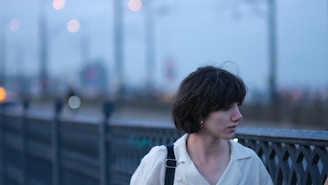A person with short dark hair stands next to a metal railing. The background is blurred, featuring soft lights and hints of distant structures.